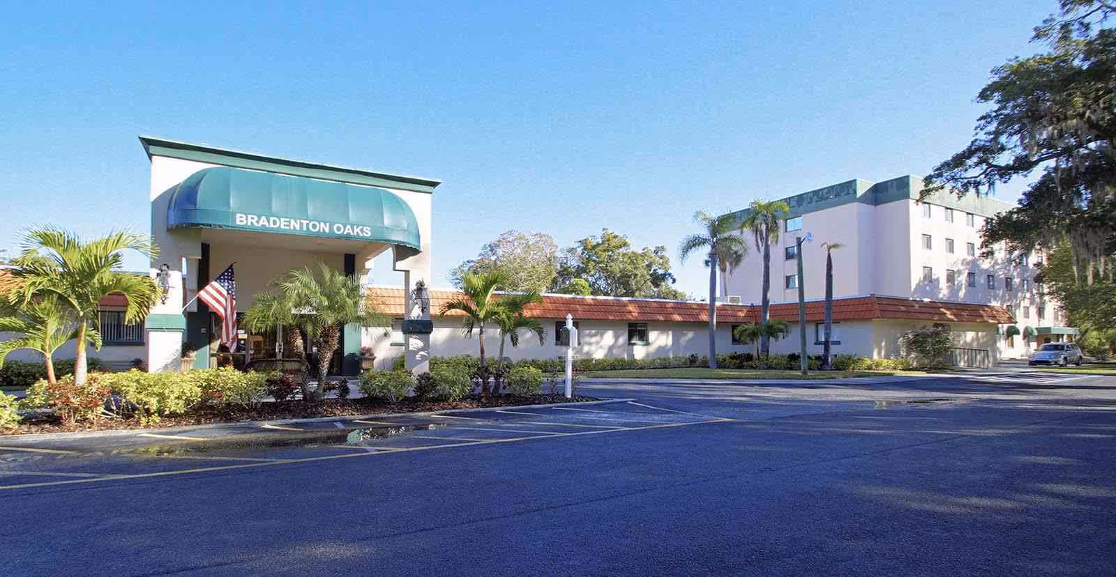 Front exterior of an assisted living building with a green awning reading "Bradenton Oaks", palm trees, and an empty parking lot.