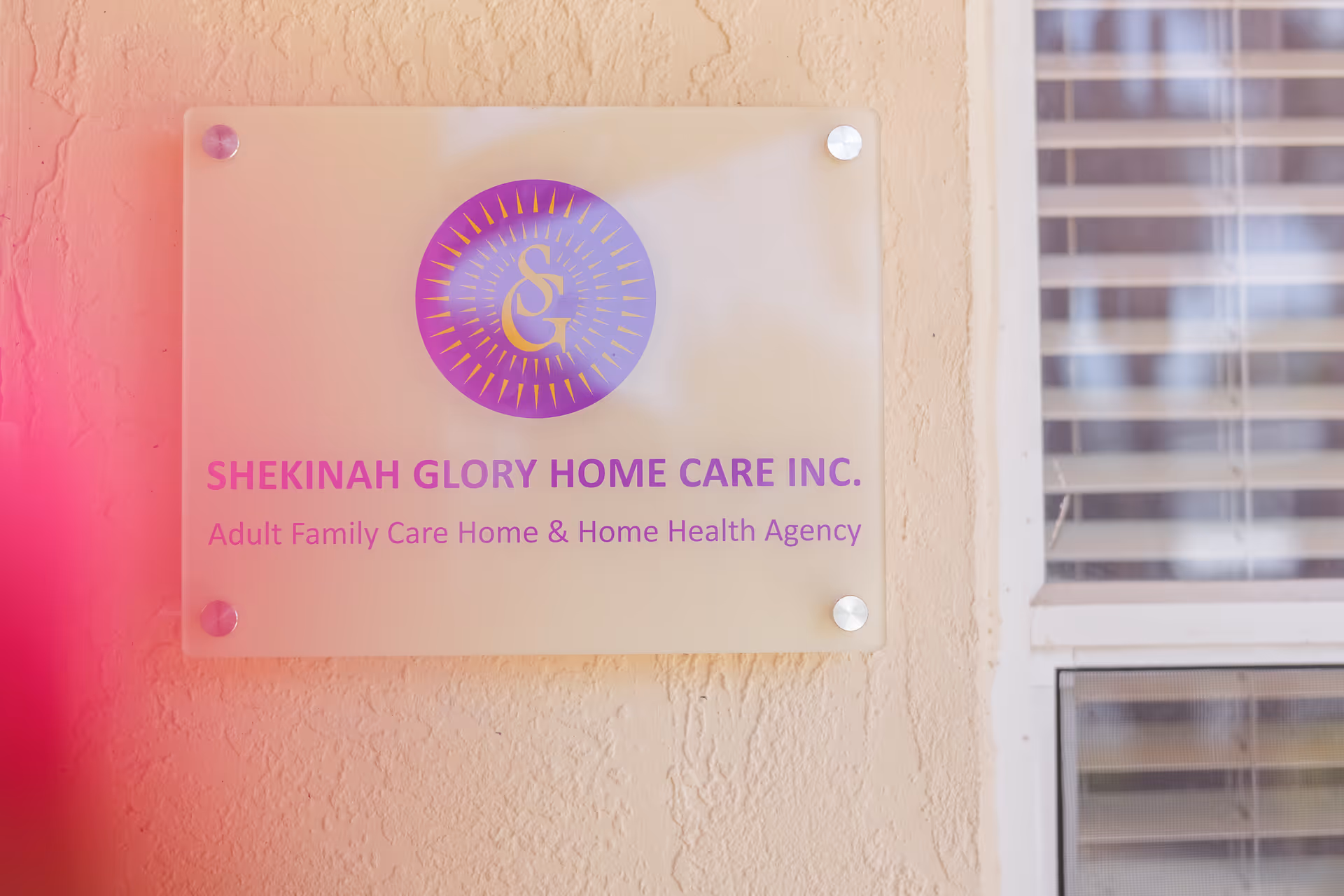 Sign for Shekinah Glory Home Care Inc. mounted on a textured exterior wall beside a window.