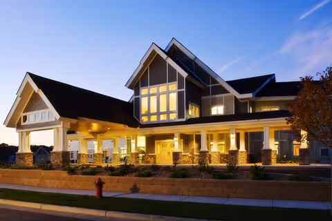 Exterior view of a large, well-lit senior living facility building at dusk with a covered entrance, multiple windows, and a landscaped area in front including a fire hydrant.
