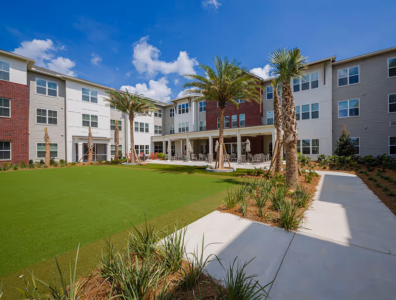Courtyard of a three-story senior living building with palm trees, a manicured lawn and patio seating under a blue sky.