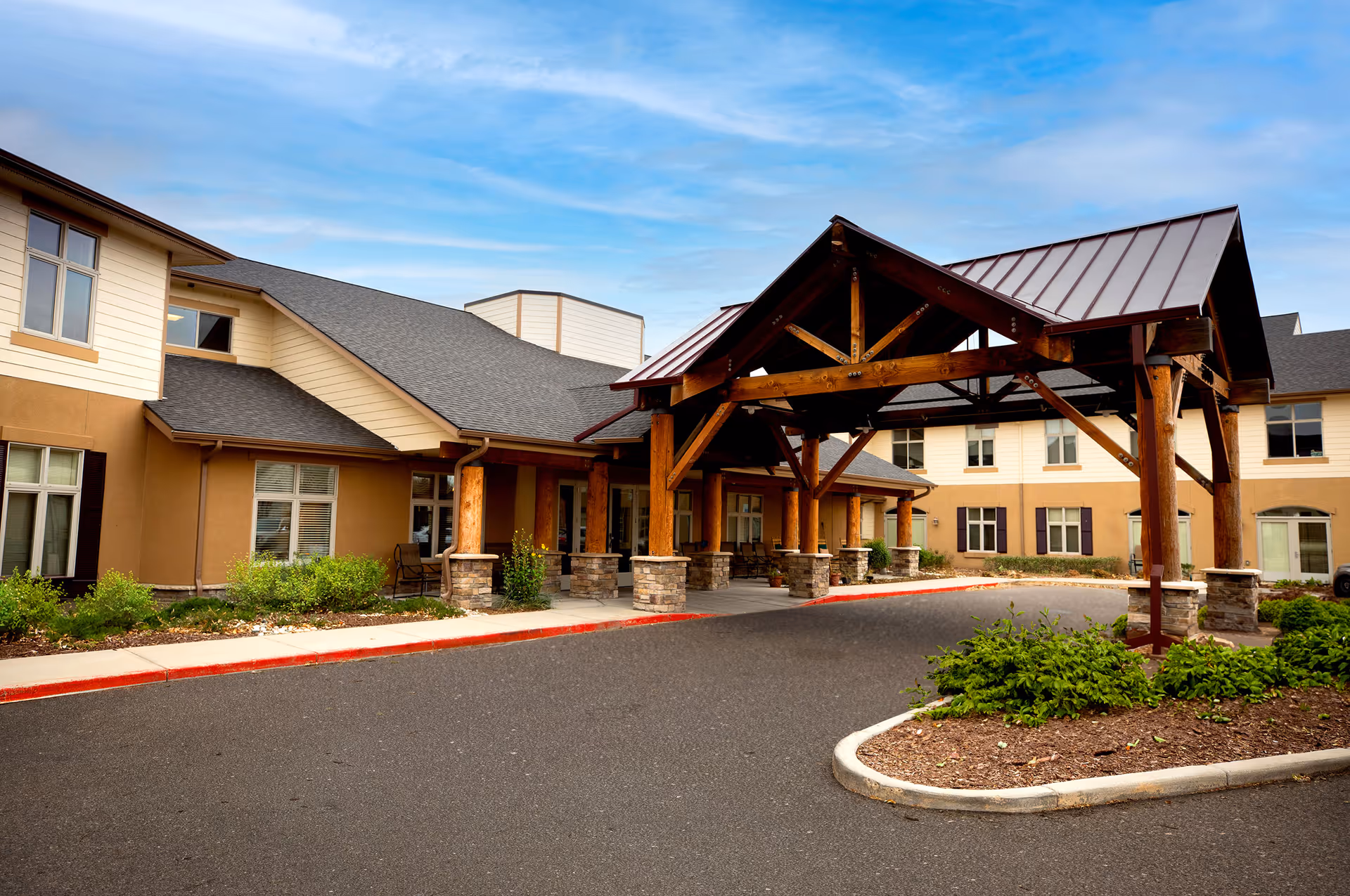 Exterior view of MeadowView of Greeley senior living facility showing the entrance with a large wooden covered driveway, beige and white building facade, windows, and landscaped greenery under a partly cloudy sky.