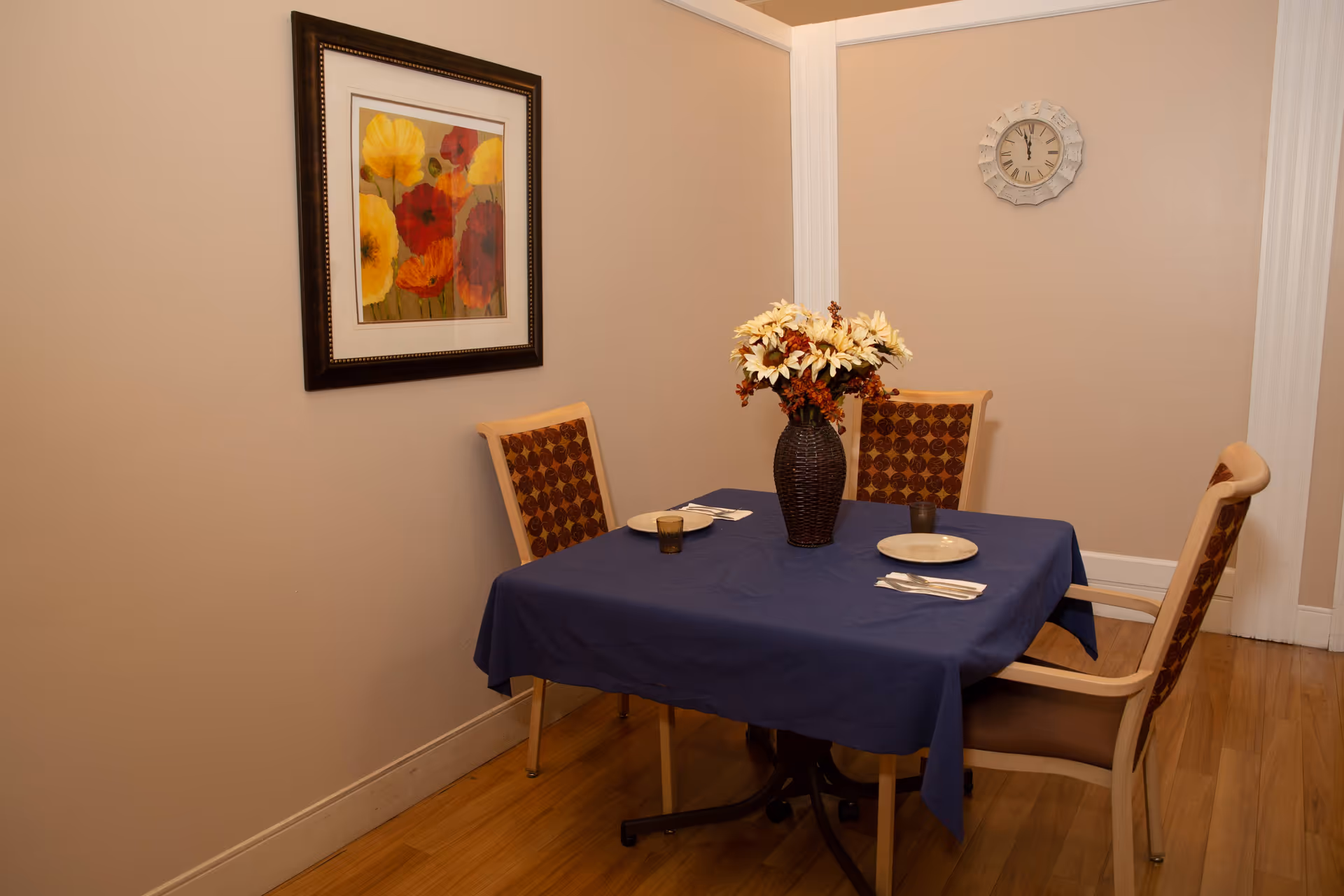 A dining table with a blue cloth, flower centerpiece, and four chairs in a beige room.