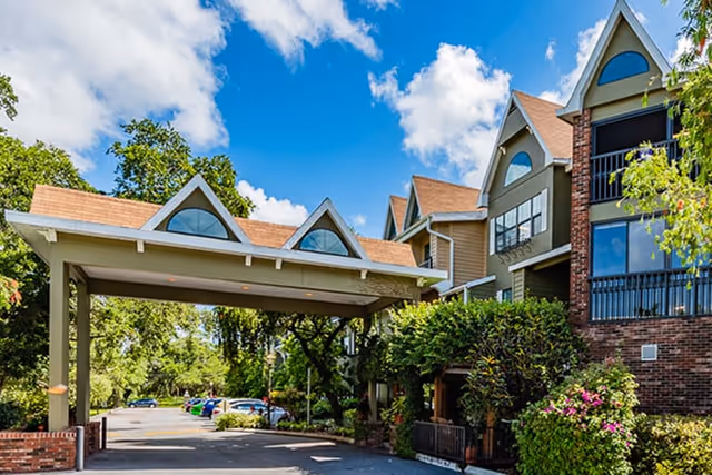 Exterior view of a senior living facility with a covered entrance, multiple peaked roof sections, brick and siding facade, surrounded by trees and greenery under a partly cloudy sky.