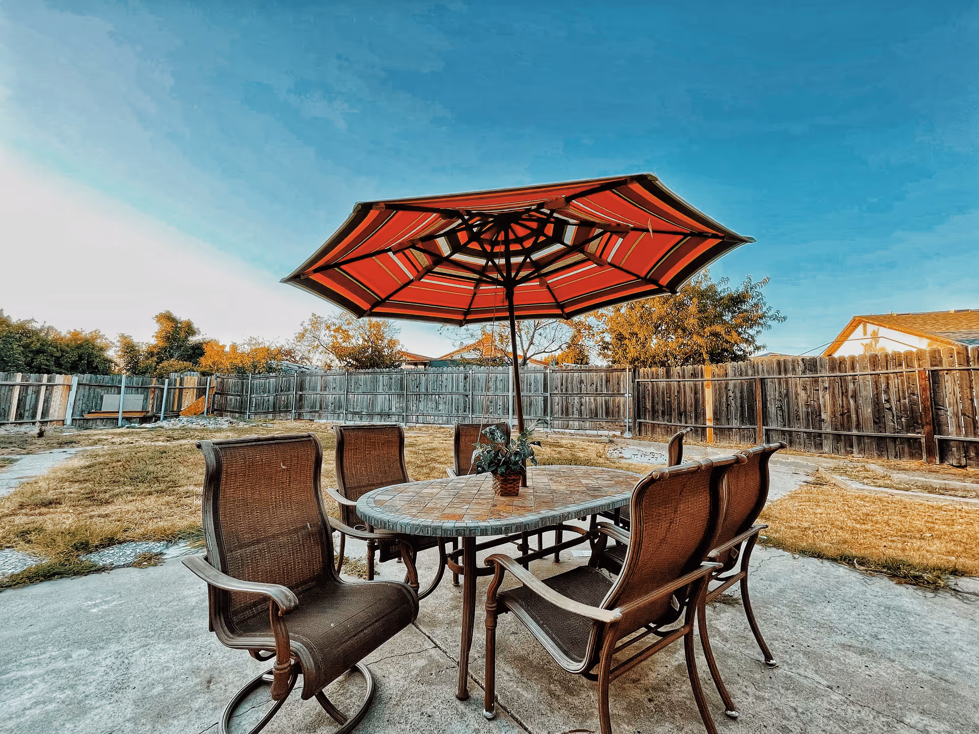 Outdoor patio area with a round table and six brown chairs arranged around it. A large red and white striped umbrella is open above the table. The patio is surrounded by a wooden fence and there is grass and trees in the background under a clear blue sky.