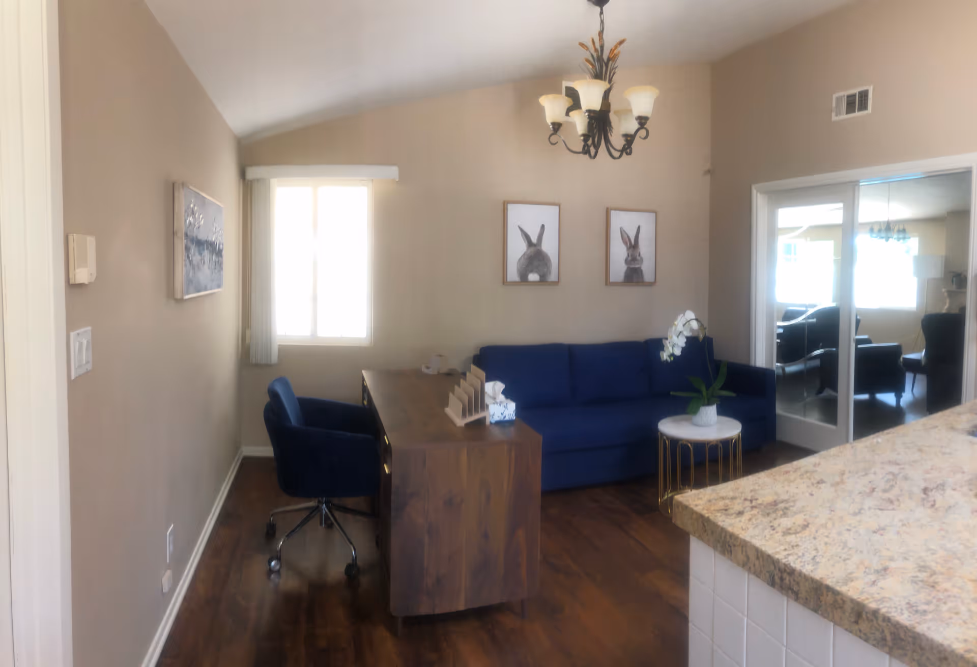 Cozy living room with blue sofa, desk, chandelier, and sliding glass doors to another room.