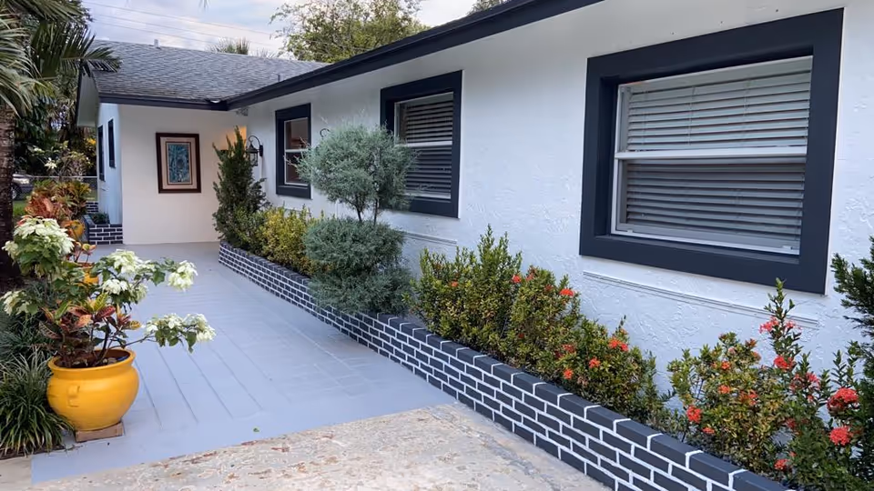 White building exterior with black window frames, a paved path, and planted shrubs