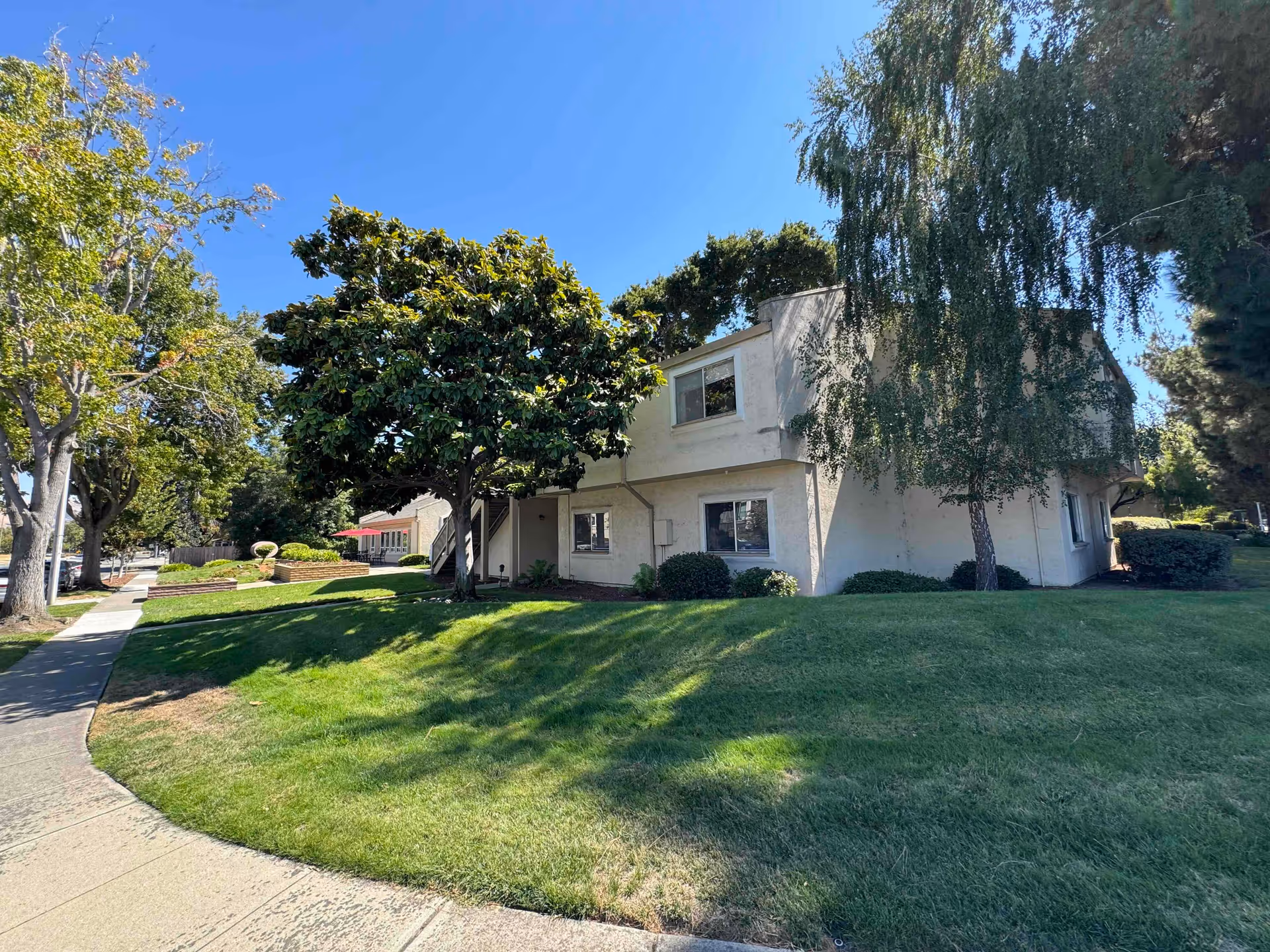 Two-story white stucco residential building with a green lawn, trees, and a sidewalk under a clear blue sky.