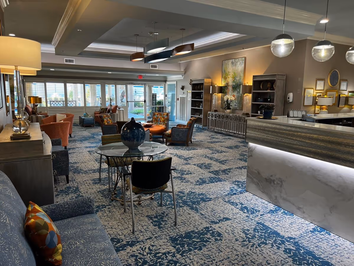 A spacious and well-lit common area in a senior living facility featuring multiple seating arrangements with armchairs and sofas, a round glass table with chairs, decorative lamps, artwork on the walls, and a reception desk with pendant lights above. Large windows with blinds allow natural light to enter the room.