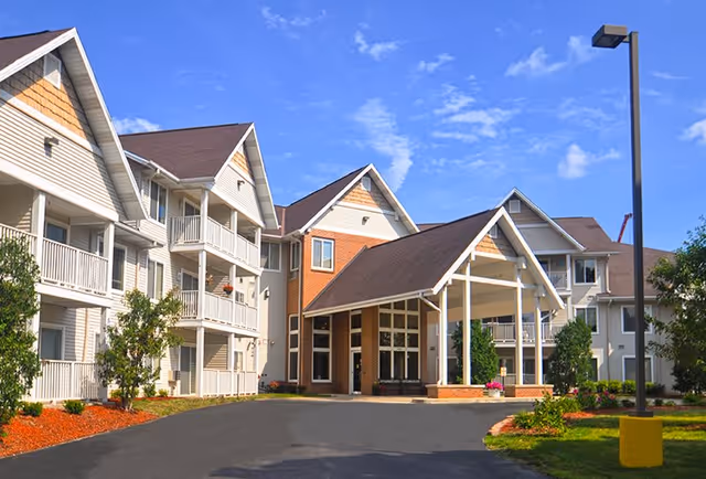 Exterior view of a senior living facility with multiple connected buildings featuring balconies, a covered entrance, landscaped greenery, and a clear blue sky.