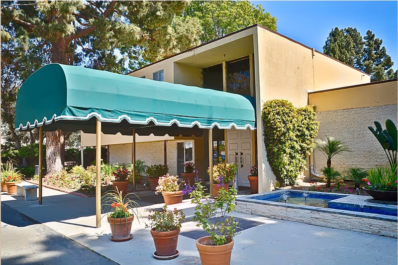 Entrance of a beige building with a green awning, potted plants, and a small fountain.
