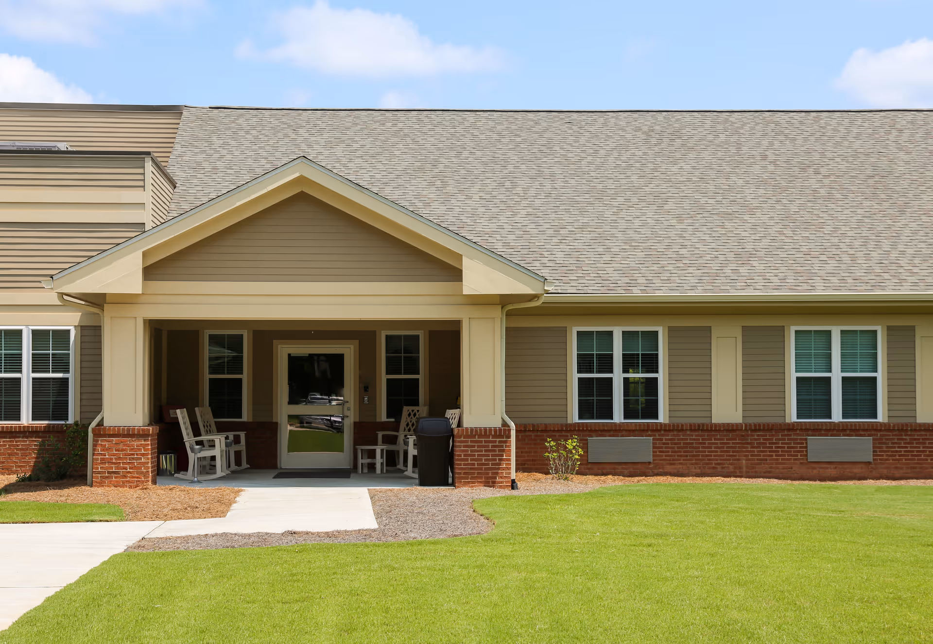 Front exterior view of a single-story building with a covered entrance porch. The building has beige siding with red brick accents, multiple windows with white trim, and a gray shingled roof. There are white rocking chairs and a small table on the porch, with a trash bin nearby. The foreground features a green lawn and a concrete walkway leading to the entrance.