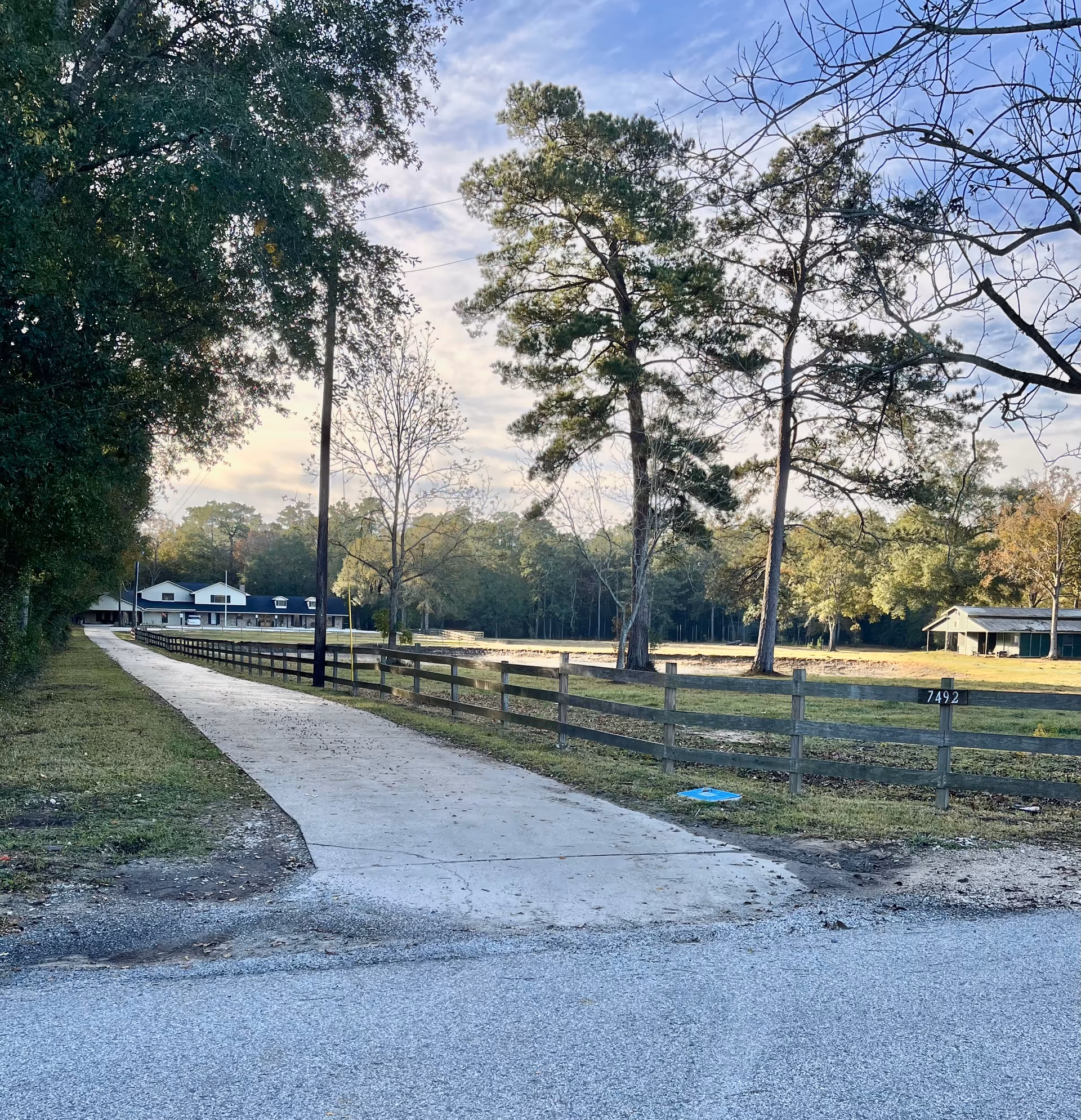 Winding concrete path through a rural campus with trees, fence, and distant buildings.