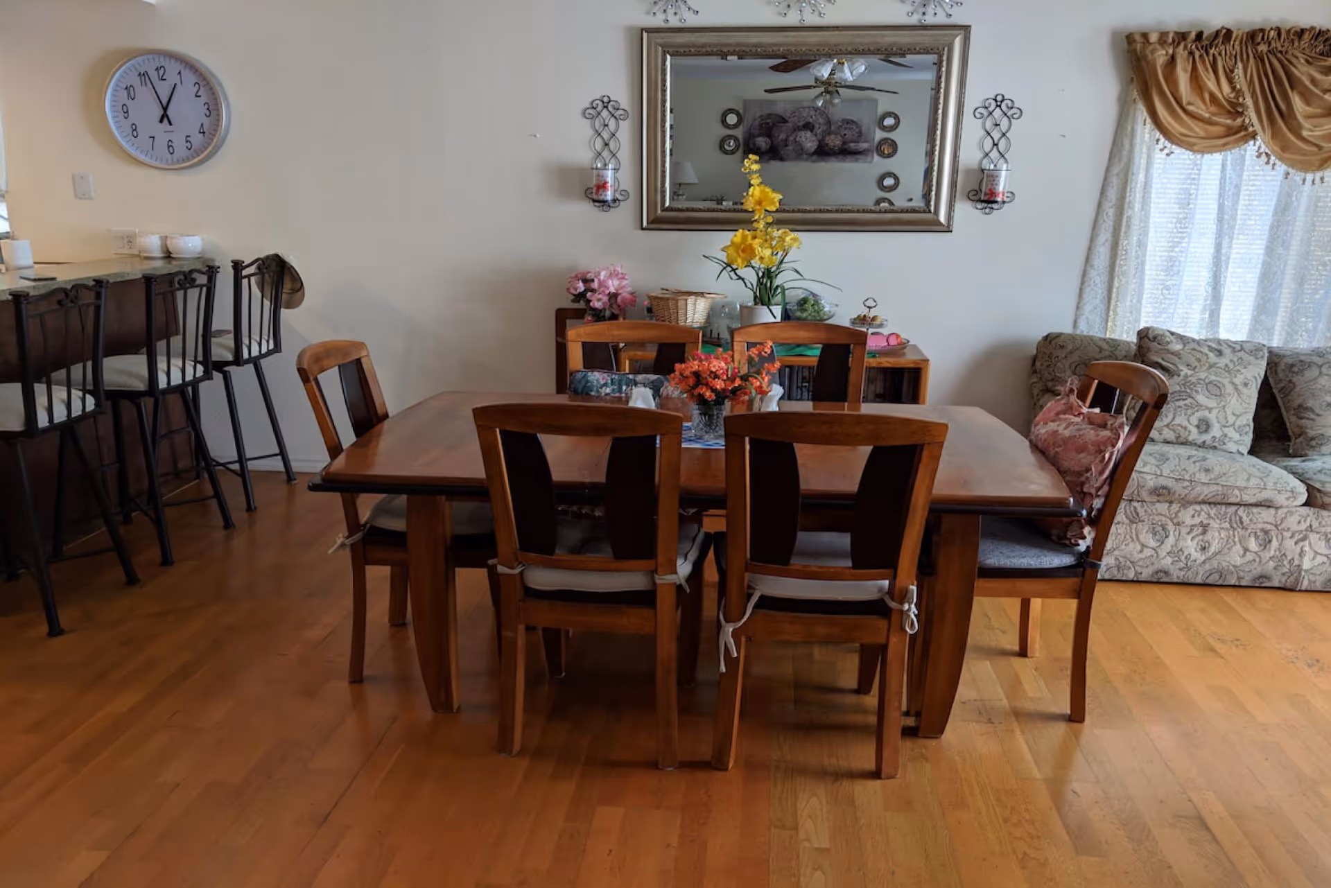 A dining room with a wooden table and six chairs, each with a cushion tied to the seat. The table has a small floral centerpiece. Behind the table is a sideboard with decorative items including flowers and a basket. A large mirror hangs on the wall above the sideboard, reflecting part of the room. To the right, there is a patterned sofa with a floral pillow and a window with sheer curtains and a gold valance. On the left side, there is a kitchen counter with three bar stools and a wall clock showing the time as 11:55.