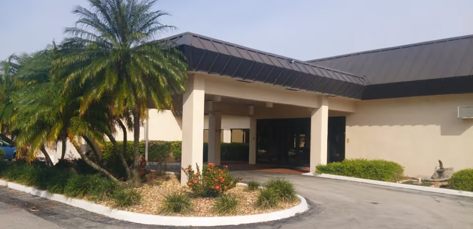Exterior view of Sunny Hills Alf - Assisted Living Facility Sebring showing the entrance with a covered driveway, palm trees, and landscaped bushes around the building.