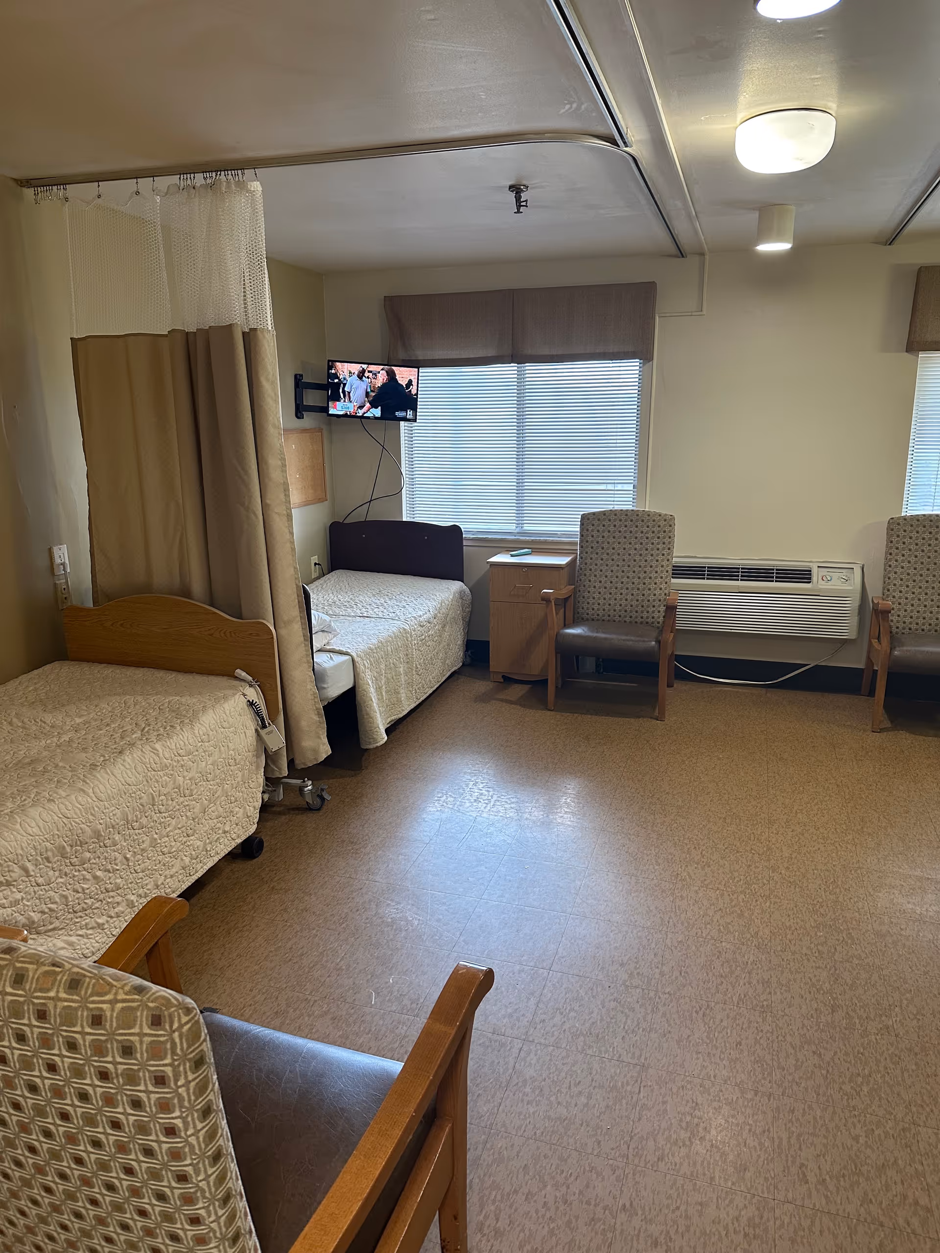Hospital patient room with two beds, privacy curtain, window blinds, chairs, and a wall-mounted TV.