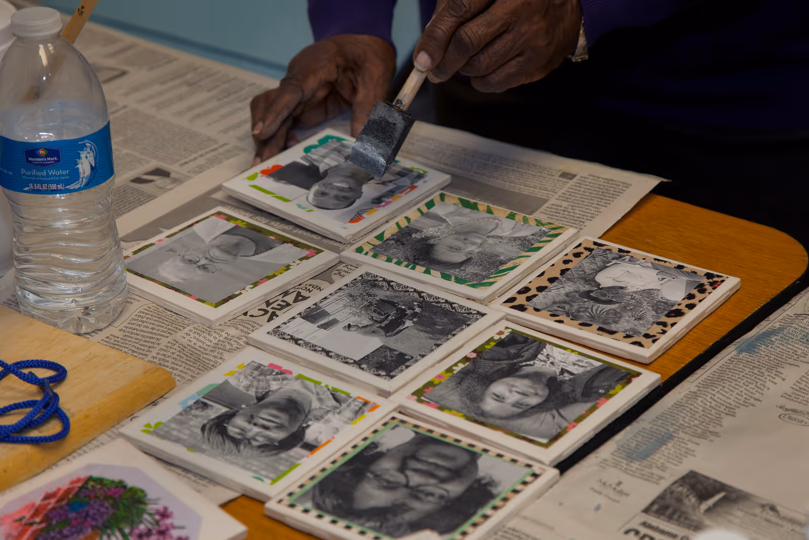 Hands apply paint to photo tiles laid on a table covered with newspaper.