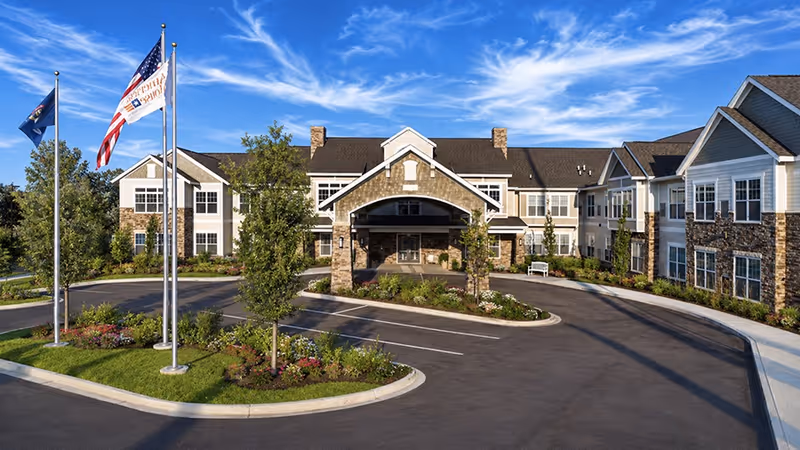 Exterior view of American House Hampton Village with a circular driveway, flags, and stone facade.