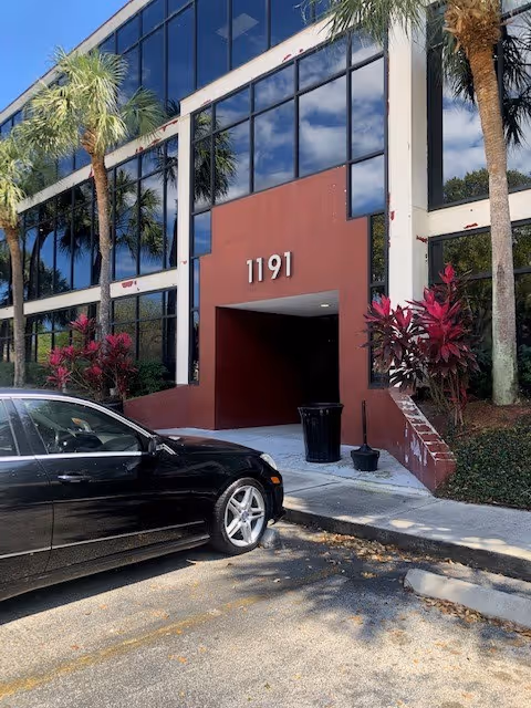 Modern glass-front building with red entrance marked 1191; black car parked outside, palm trees nearby.