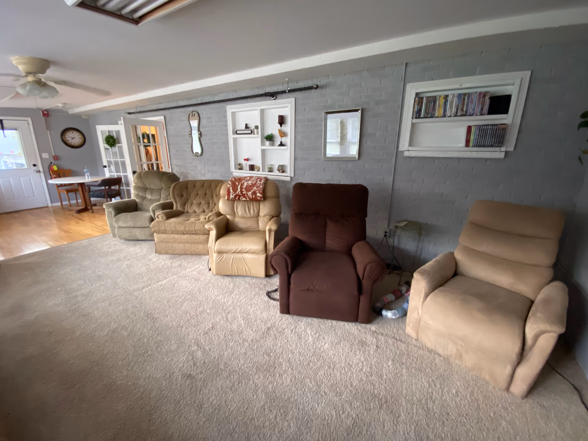 A cozy living room area with five recliner chairs lined up against a gray brick wall. The wall features built-in shelves with decorative items and a small bookshelf with DVDs. The room has beige carpet flooring and a wooden floor area near a door with glass panels. A ceiling fan and a wall clock are also visible.