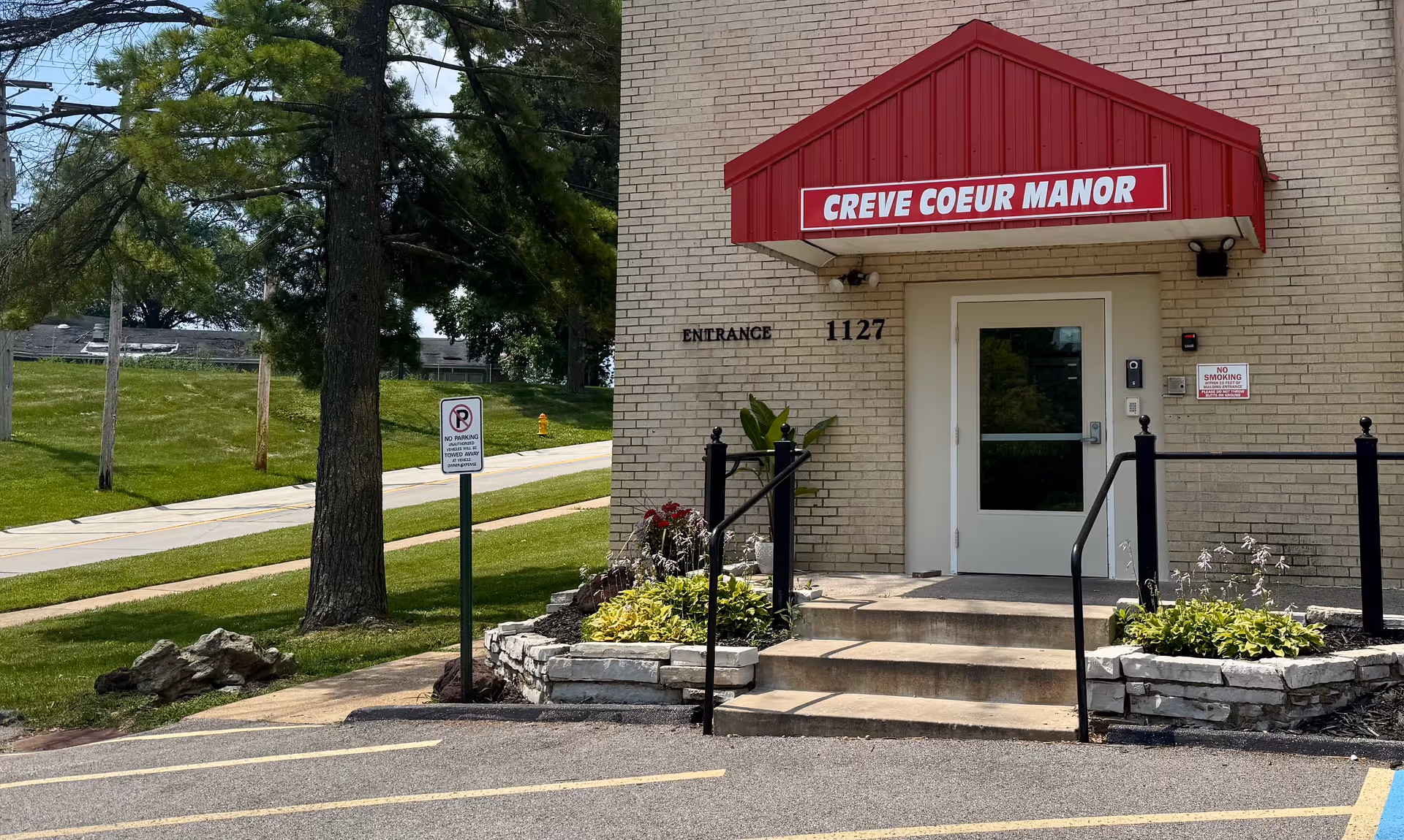 Entrance to Creve Coeur Manor, a beige brick building with a red awning above the door displaying the facility name. There are steps leading up to the door with black handrails on either side, small landscaped areas with plants and flowers, and a no parking sign on a pole near the sidewalk. Green grass and trees are visible in the background.
