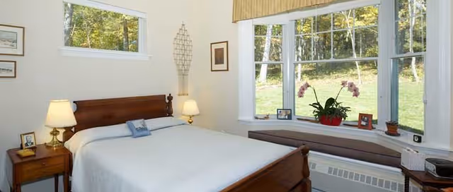 A cozy bedroom with a wooden bed frame and white bedding. There are two bedside tables with lamps and framed photos. A large bay window with a cushioned seat overlooks a green outdoor area with trees. The room has light-colored walls and simple decor.