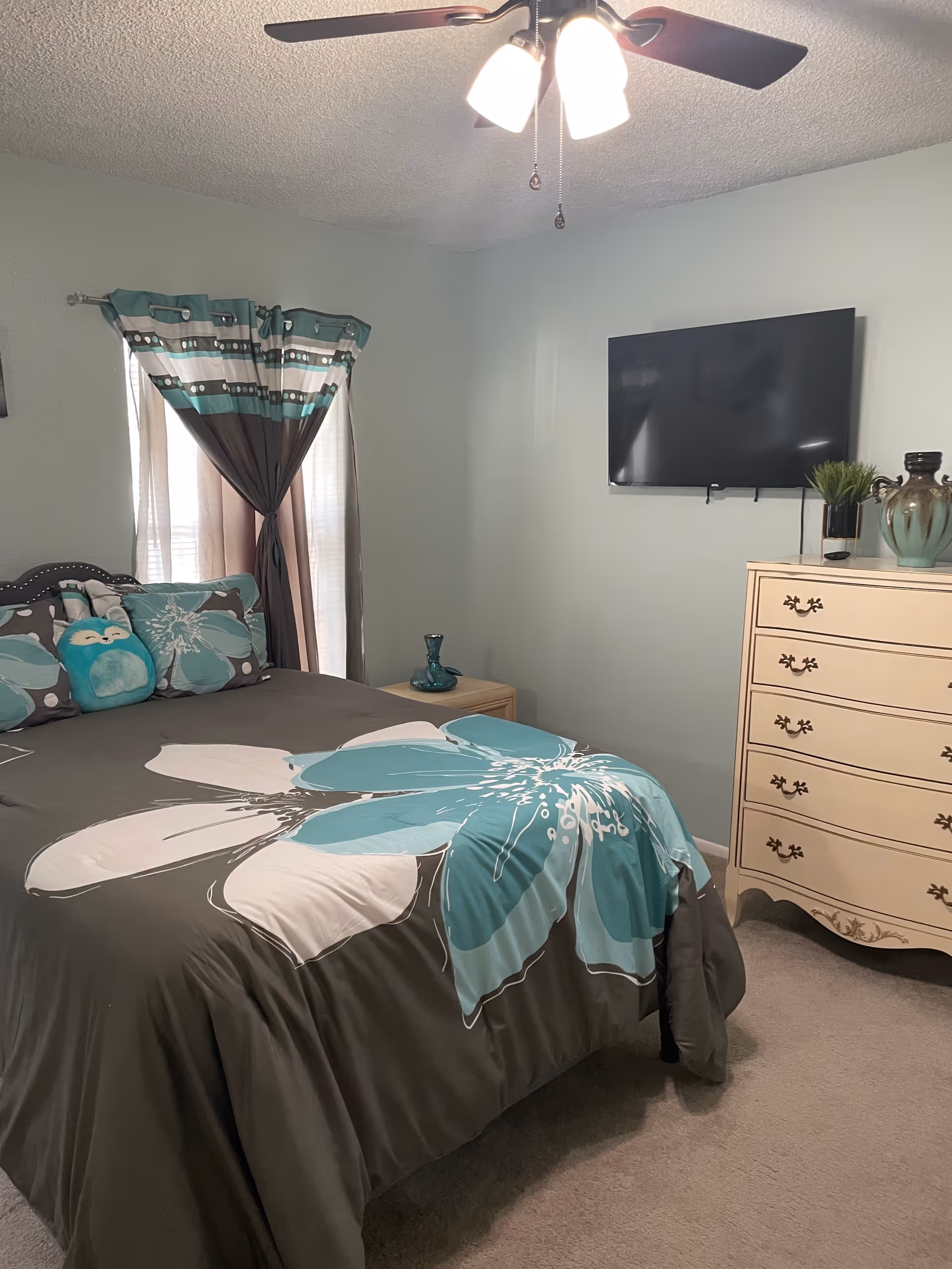 Bedroom in teal/gray tones with bed, dresser, wall-mounted TV, and ceiling fan.