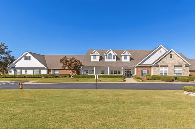 Front exterior view of a single-story senior living facility building with a well-maintained lawn, shrubs, and a clear blue sky.