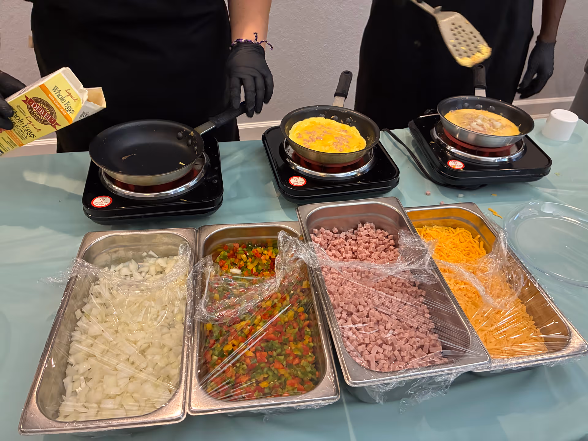 Staff wearing gloves cook on stoves; trays hold chopped onions, peppers, ham, cheese.