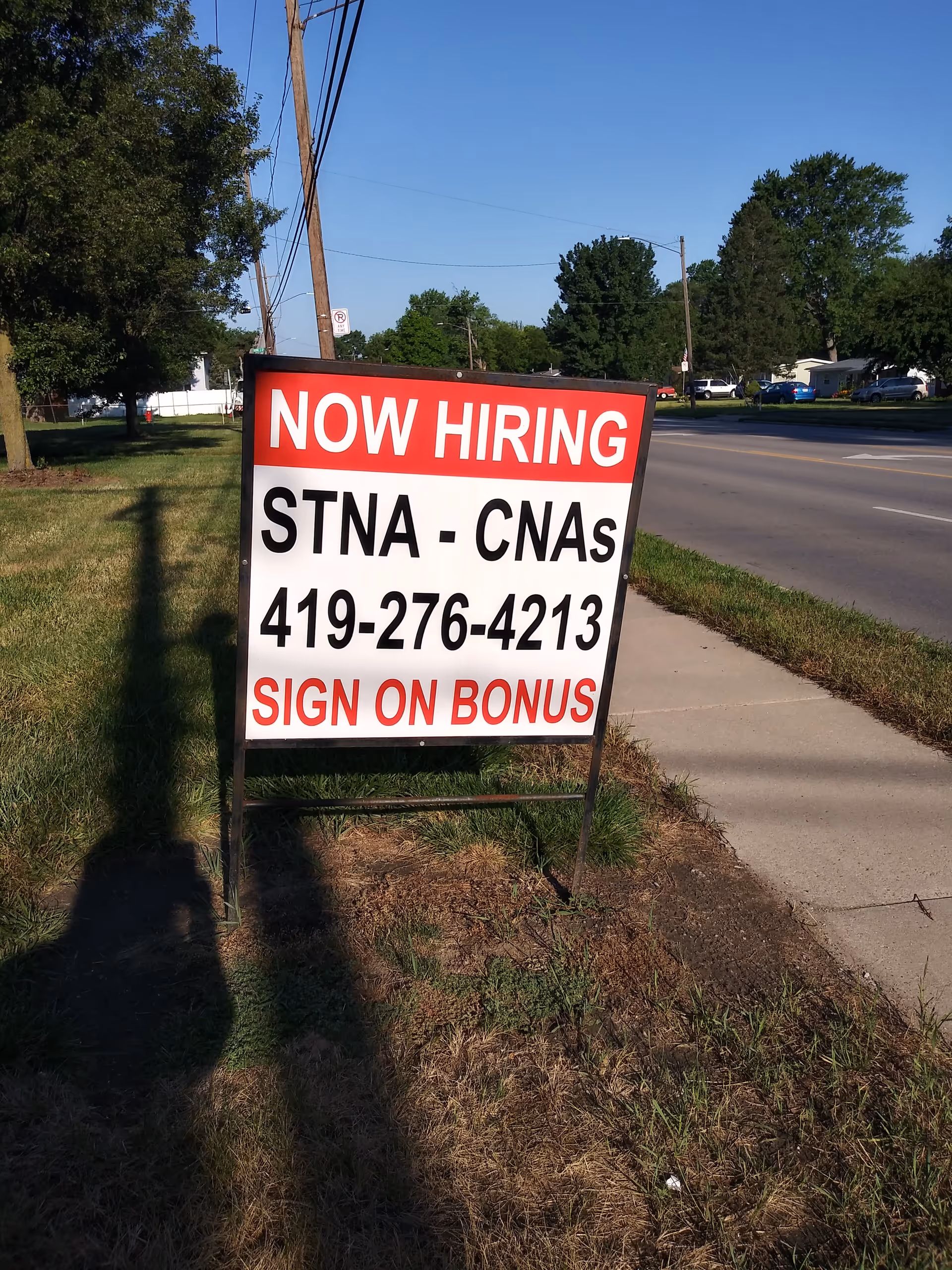 A roadside lawn displays a large NOW HIRING sign for STNA-CNAs with a phone number.