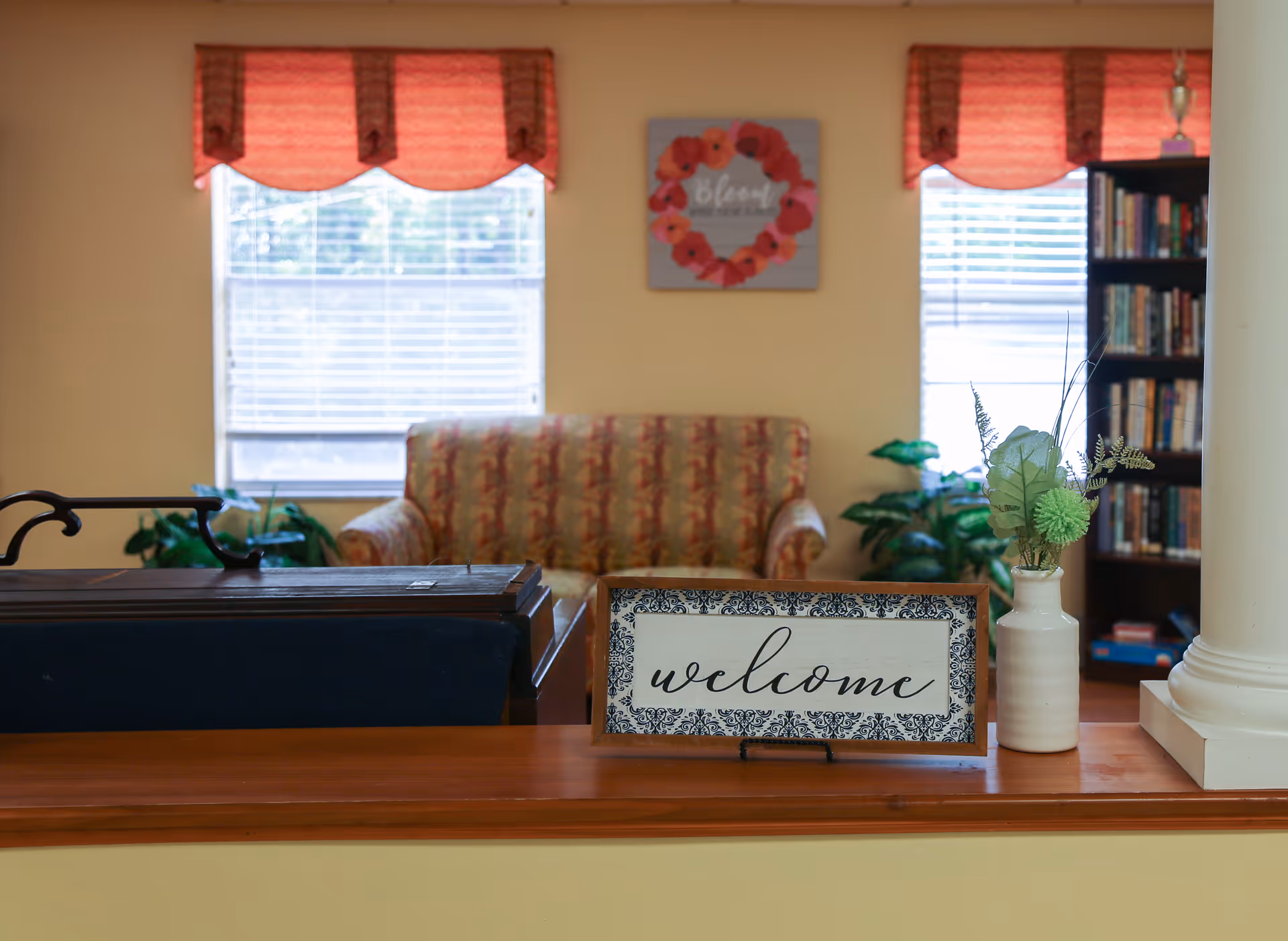 A cozy living room area with a patterned sofa in front of two windows with red valances. A framed sign with the word 'welcome' and a white vase with green artificial flowers are placed on a wooden ledge in the foreground. A bookshelf filled with books is visible on the right side of the room.
