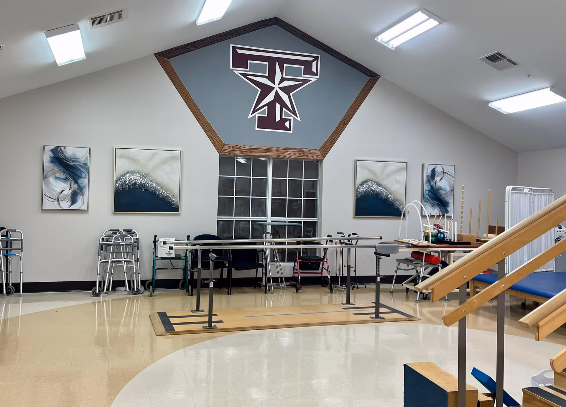 Interior of a rehabilitation therapy room with parallel bars for walking exercises, several walkers lined up against the wall, a set of stairs with handrails for physical therapy, and abstract artwork on the walls. The room has bright overhead lighting and a large logo featuring a stylized 'T' with a star above a window.