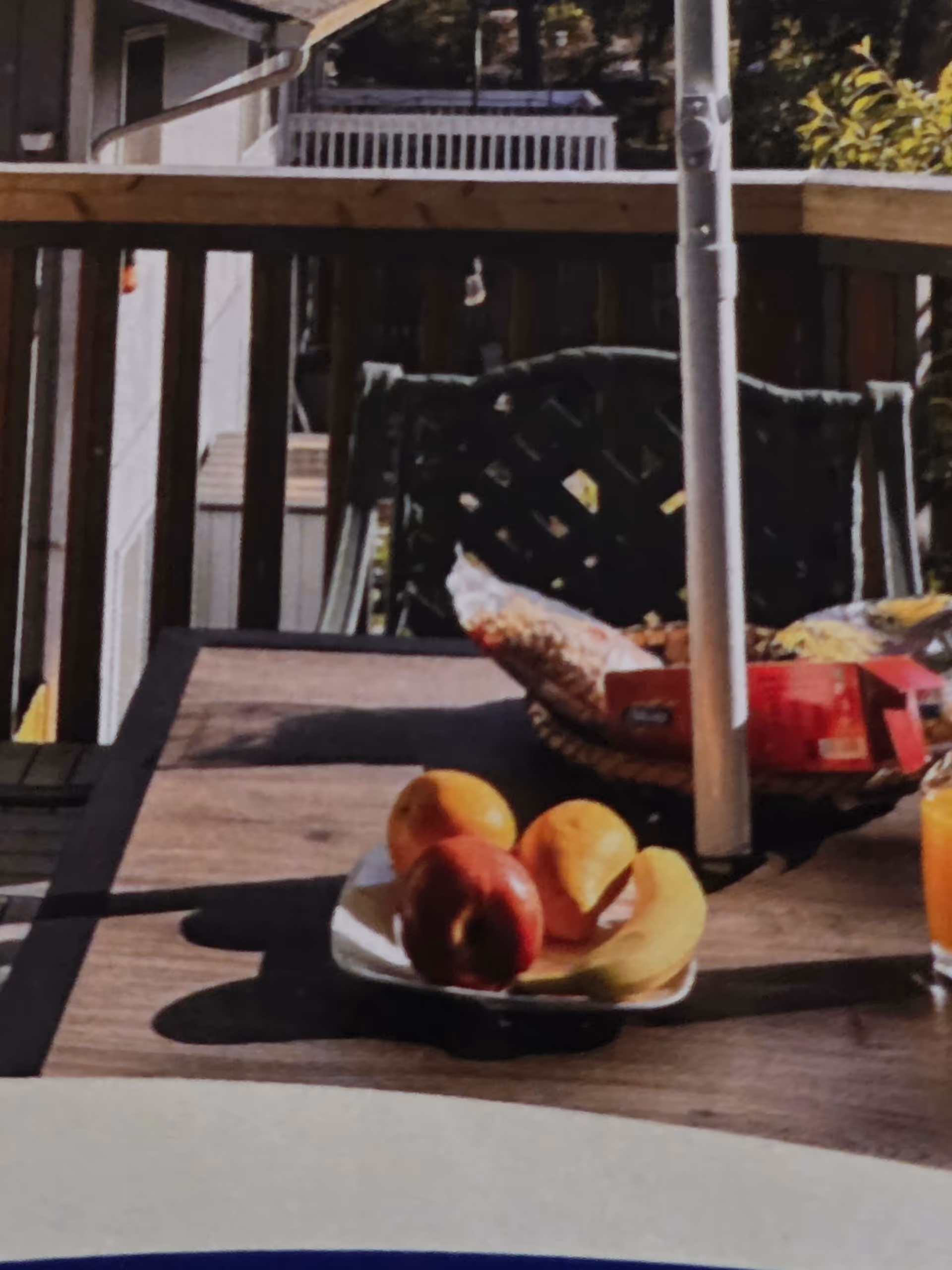 Outdoor wooden deck table with fruit plate and orange juice, railing in background.