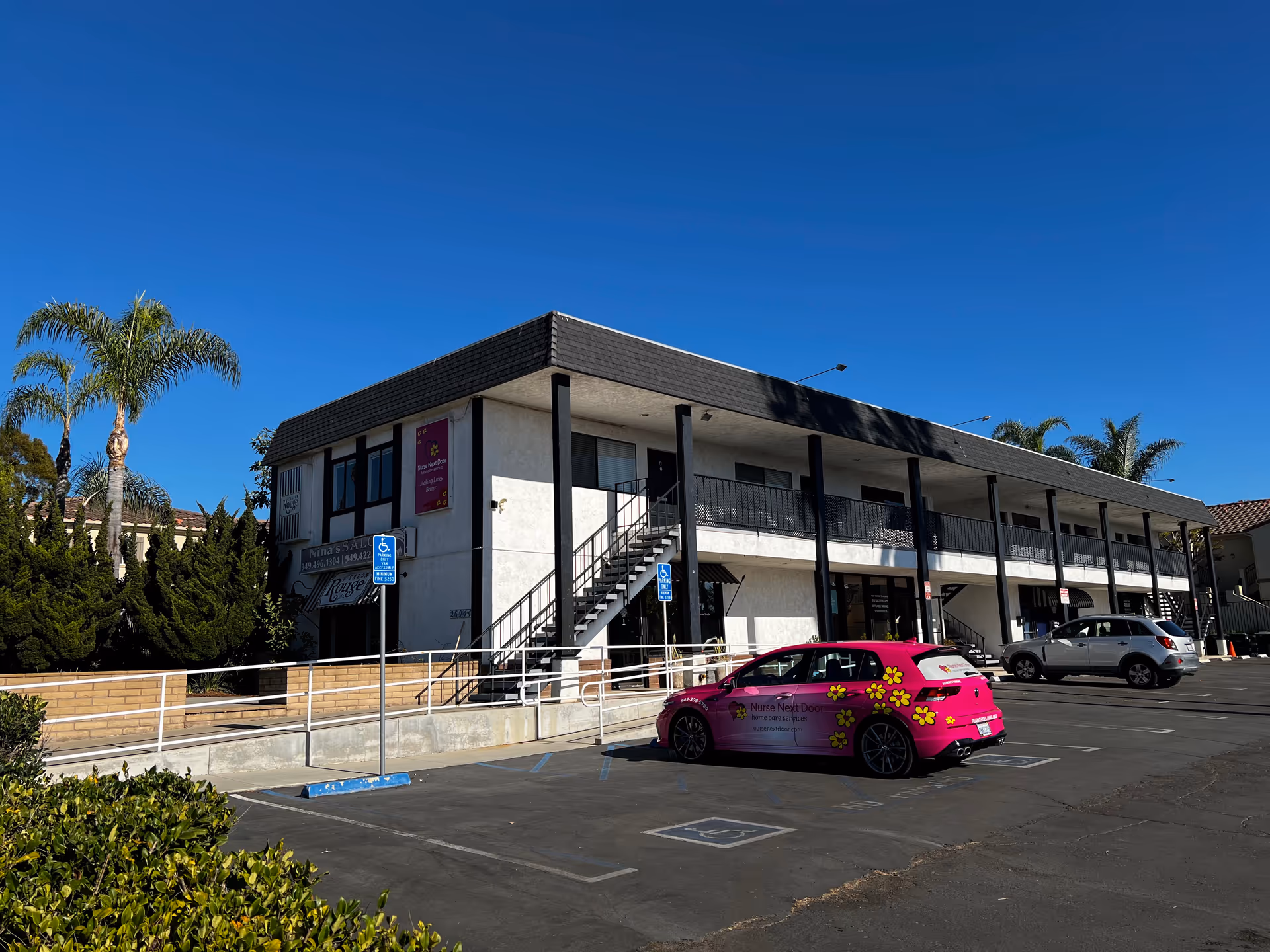 Two-story building with a parking lot, pink van, and palm trees under blue sky.