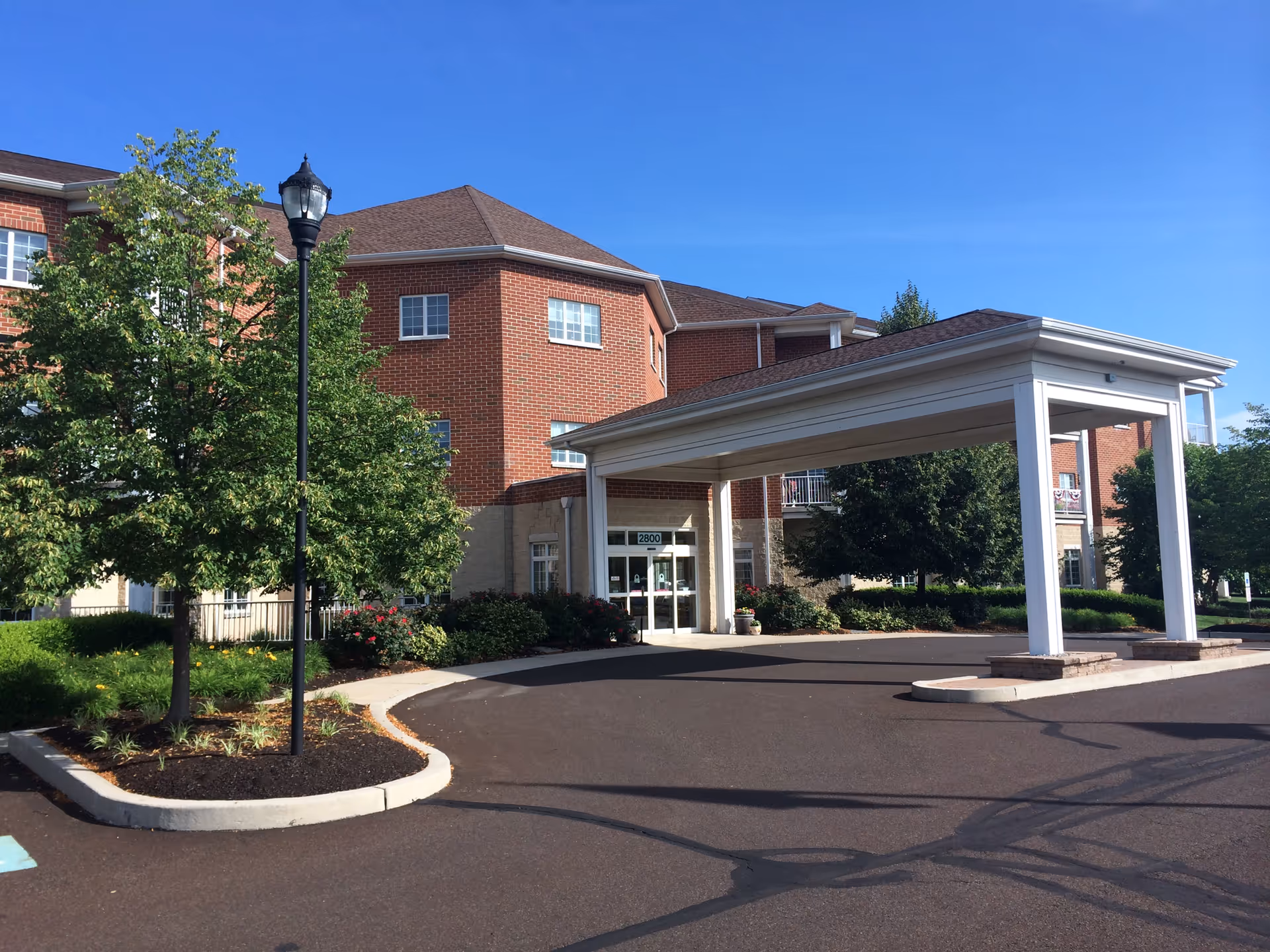 Brick senior living building with a covered entrance and manicured landscaping under a clear sky.