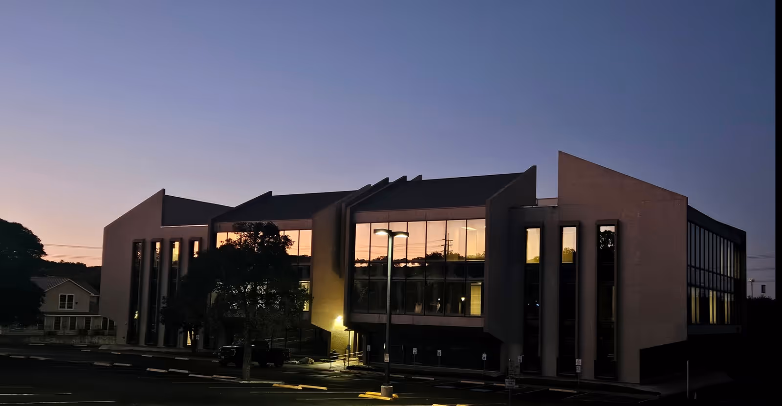 Modern two-story building with large glass windows at dusk and a parking lot