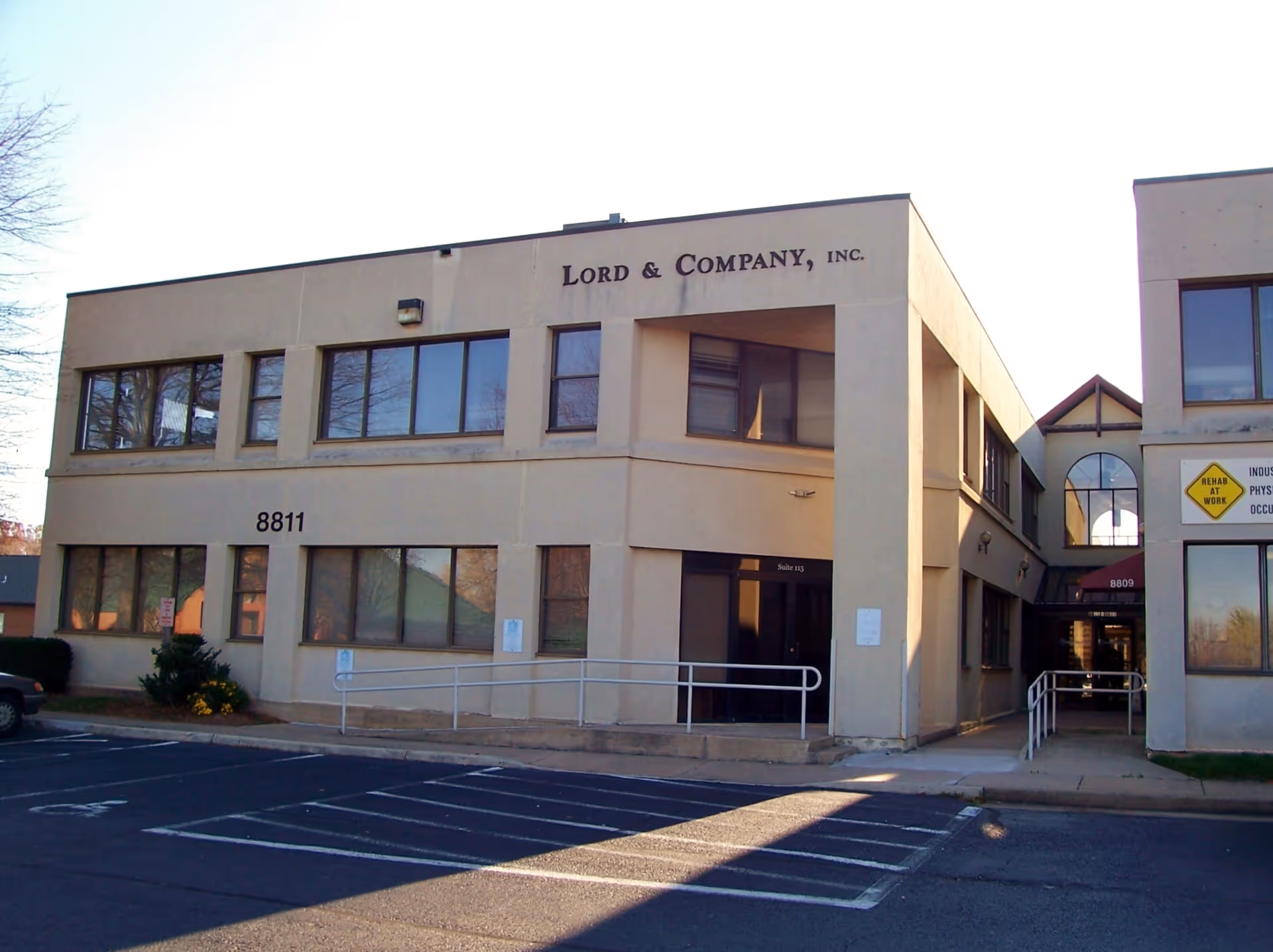 Two-story beige commercial building with large windows and a parking lot in front.