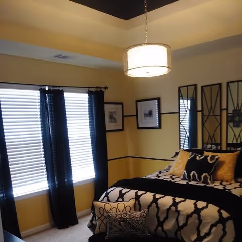 A decorated bedroom with patterned black-and-white bedding, yellow accent pillows, black curtains over a window, framed wall art, and a hanging ceiling light.