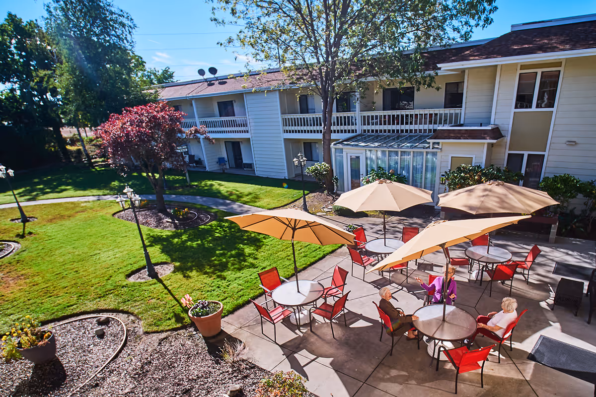 Outdoor patio area at Northridge Senior Living with several round tables, red chairs, and large beige umbrellas. Three elderly women are seated and conversing under the umbrellas. The patio is adjacent to a two-story building with balconies and surrounded by green grass, trees, and landscaped garden beds.