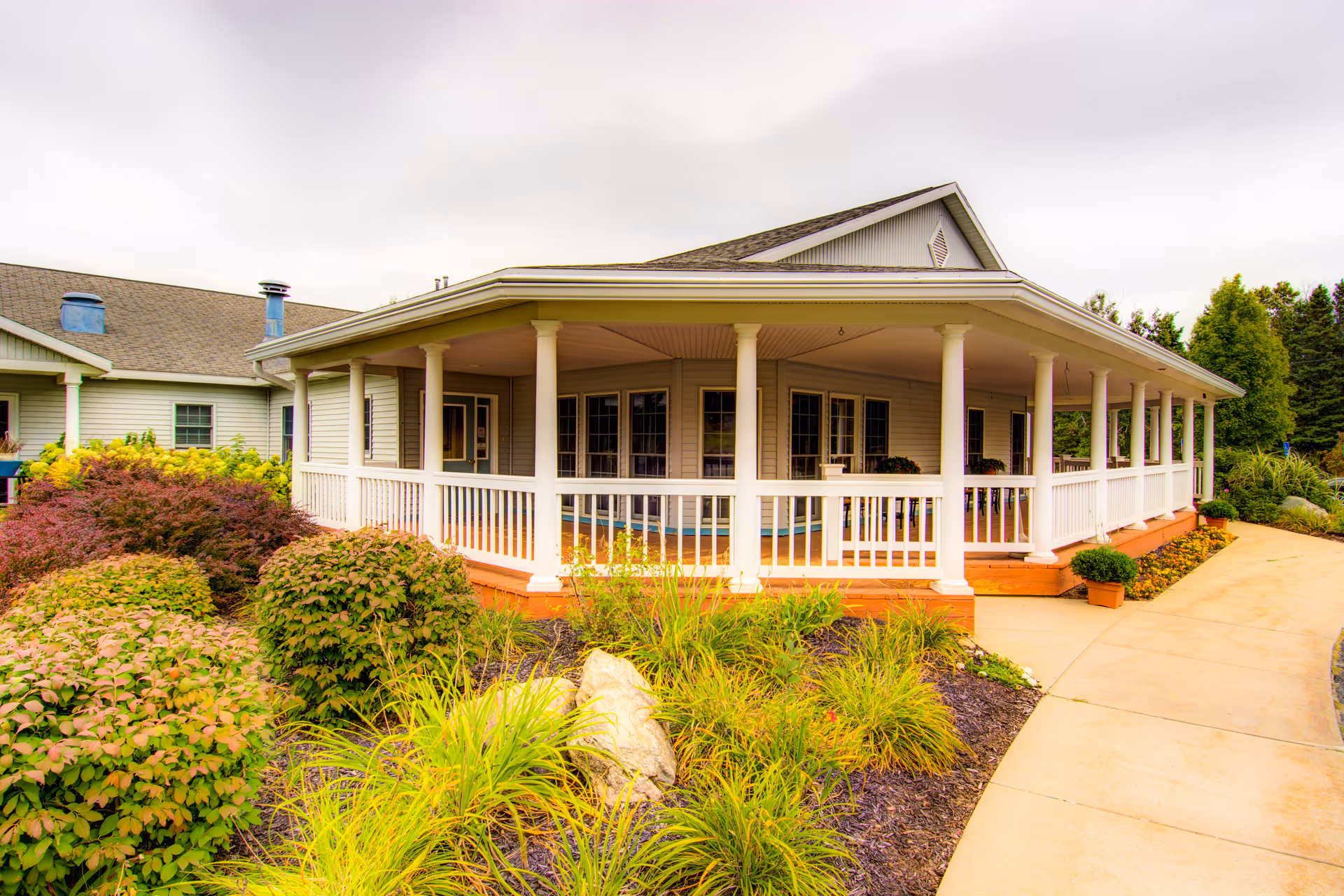 Front exterior of a single-story building with a wraparound covered porch, white railings, and landscaped shrubs and walkway.