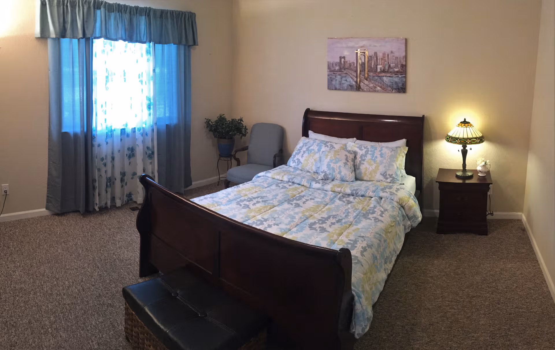 A neatly made bedroom with a wooden bed, nightstand, lamp, window, and seating.