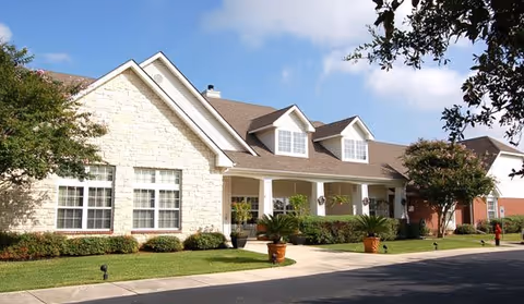 Low one-story stone and siding senior living building with dormer windows, a covered front porch and landscaped lawn under a blue sky.