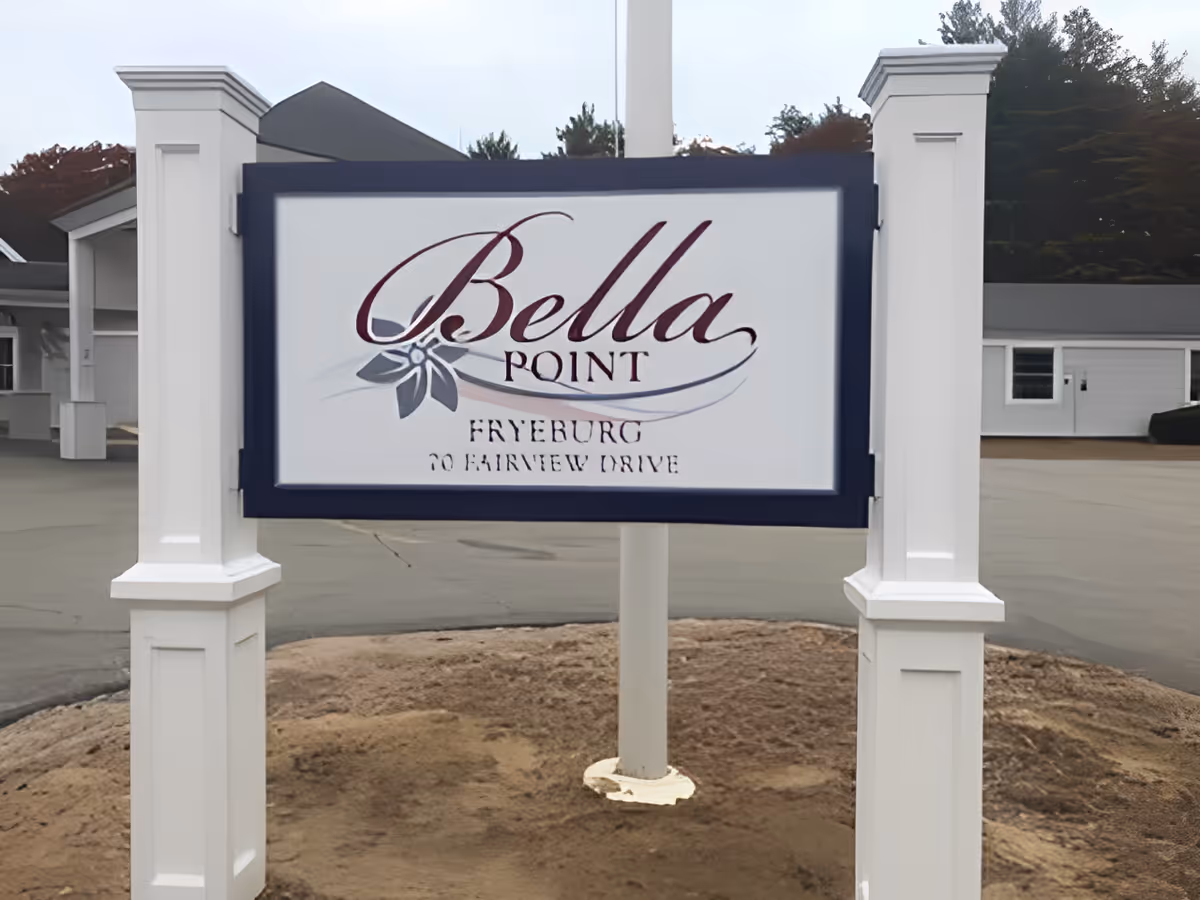 A white and navy blue sign for Bella Point Fryeburg, located at 70 Fairview Drive, mounted on two white posts with a building and paved area in the background.