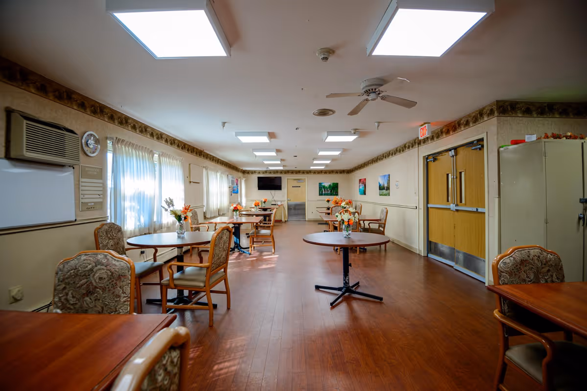 A spacious dining room in a nursing and rehabilitation center with several round and rectangular wooden tables, each adorned with a vase of flowers. The room has wooden flooring, beige walls with a decorative border near the ceiling, and multiple windows with sheer white curtains allowing natural light to enter. Ceiling lights and a ceiling fan are visible, along with a wall-mounted TV and double wooden doors at the far end.