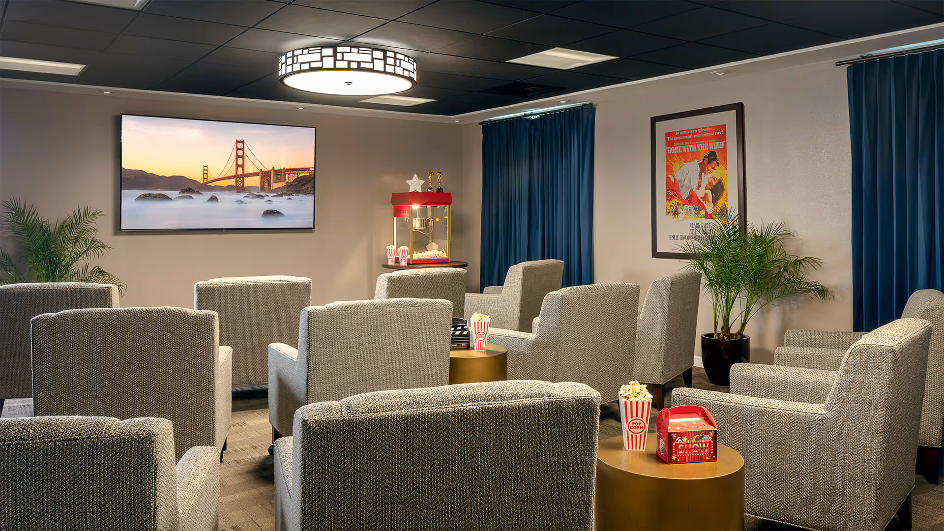A cozy movie room with multiple gray upholstered armchairs arranged in rows facing a large flat-screen TV mounted on the wall displaying an image of the Golden Gate Bridge. There is a red popcorn machine on a stand near the back wall, popcorn containers on small round tables between the chairs, blue curtains on the windows, potted plants, and a framed movie poster on the wall.
