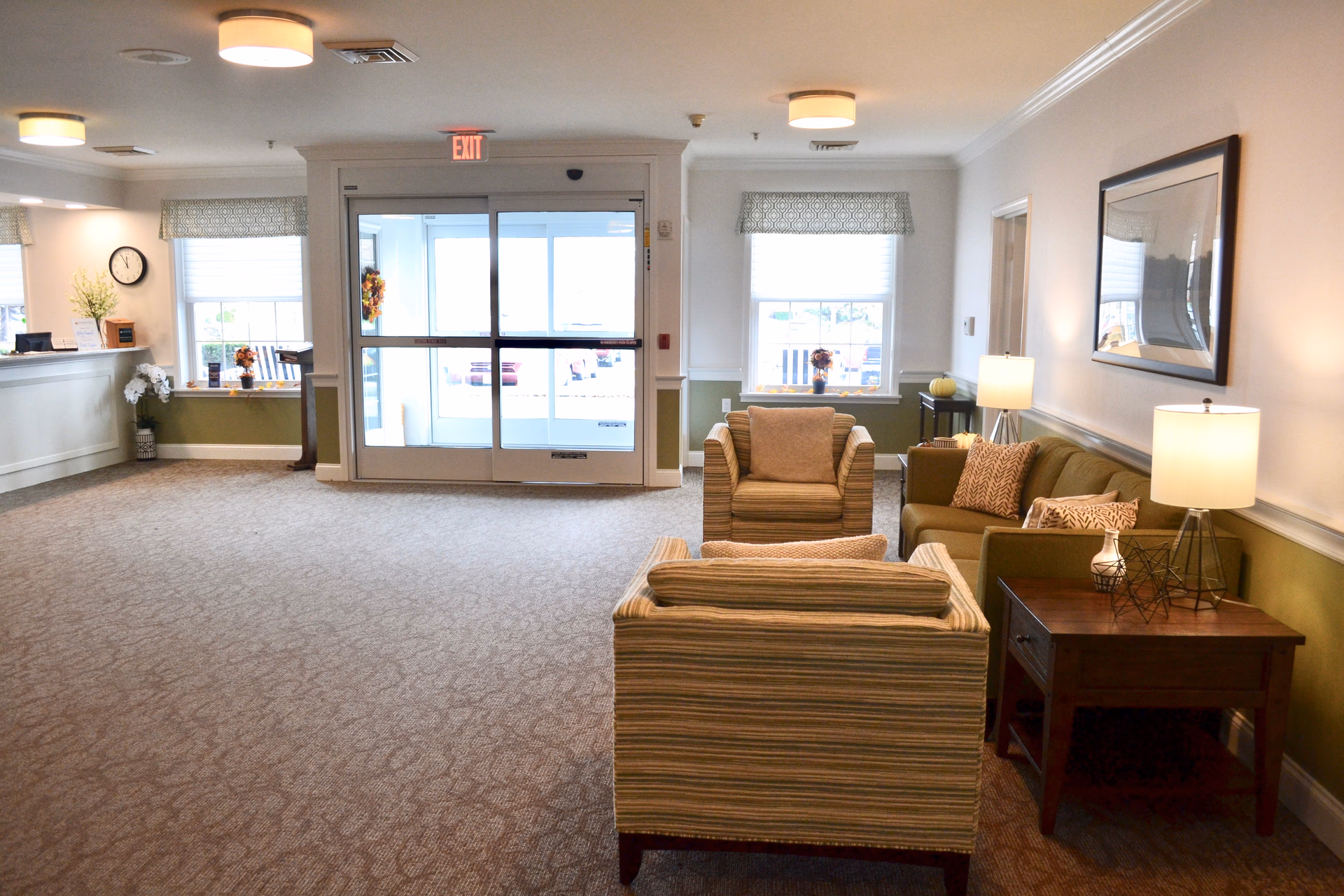 A bright and welcoming senior living facility lobby with a seating area featuring a sofa, two armchairs, and a wooden side table with a lamp. The room has carpeted floors, large windows with patterned valances, and a glass automatic door leading outside. The reception desk is visible on the left side with a clock and decorative plants.