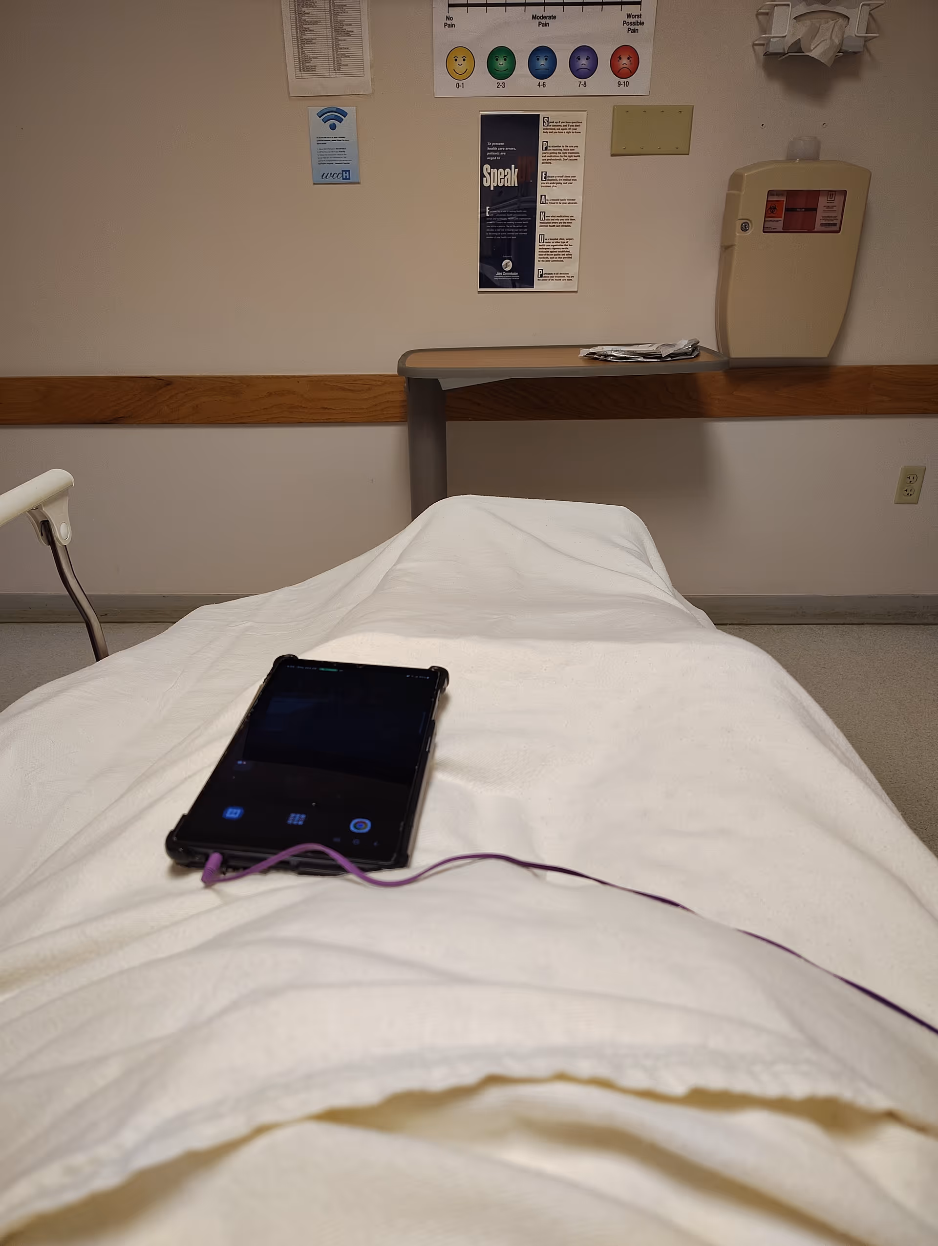 Hospital bed with white sheets, a smartphone and purple headphones resting on it.