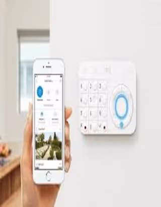 Resident uses smartphone app next to wall-mounted alarm keypad in interior.