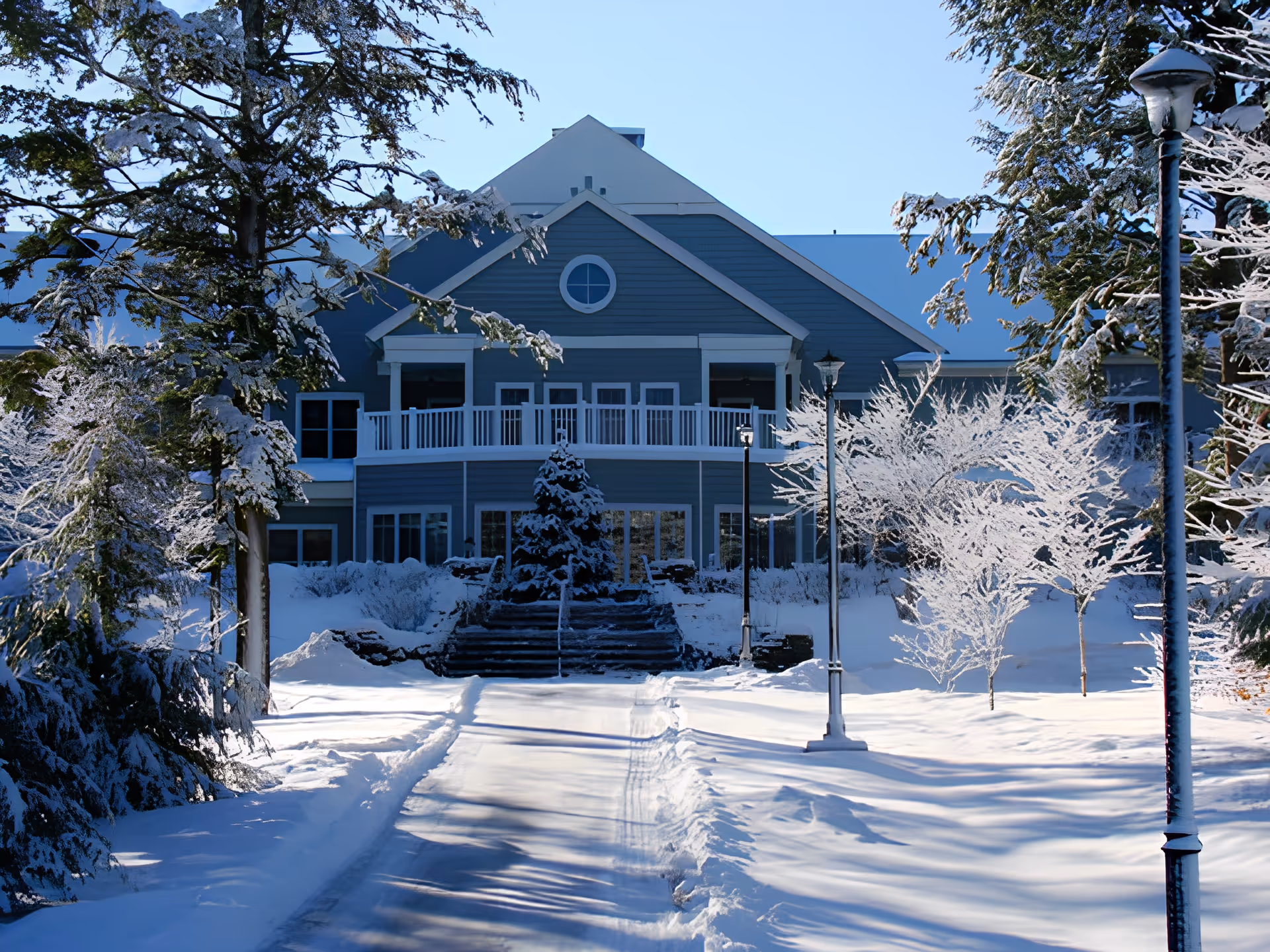 A large blue building with white trim surrounded by snow-covered trees and ground. A cleared pathway leads up to the building's entrance, which has a balcony and several windows. The scene is bright with clear blue sky and sunlight casting shadows on the snow.
