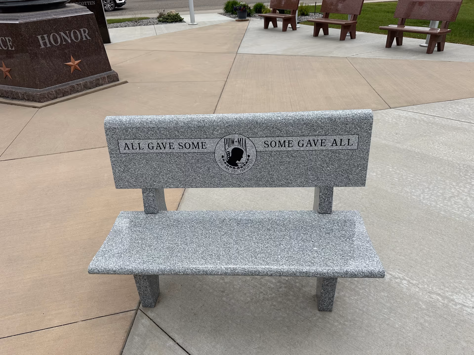 Granite outdoor memorial bench with inscription 'All Gave Some, Some Gave All' in plaza.