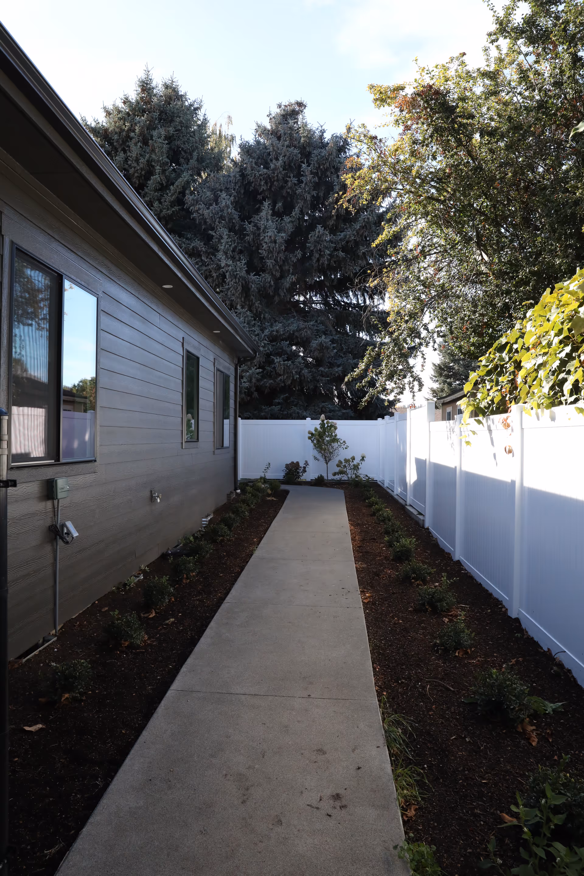 Narrow concrete walkway beside a facility's exterior, bordered by shrubs and a white privacy fence.